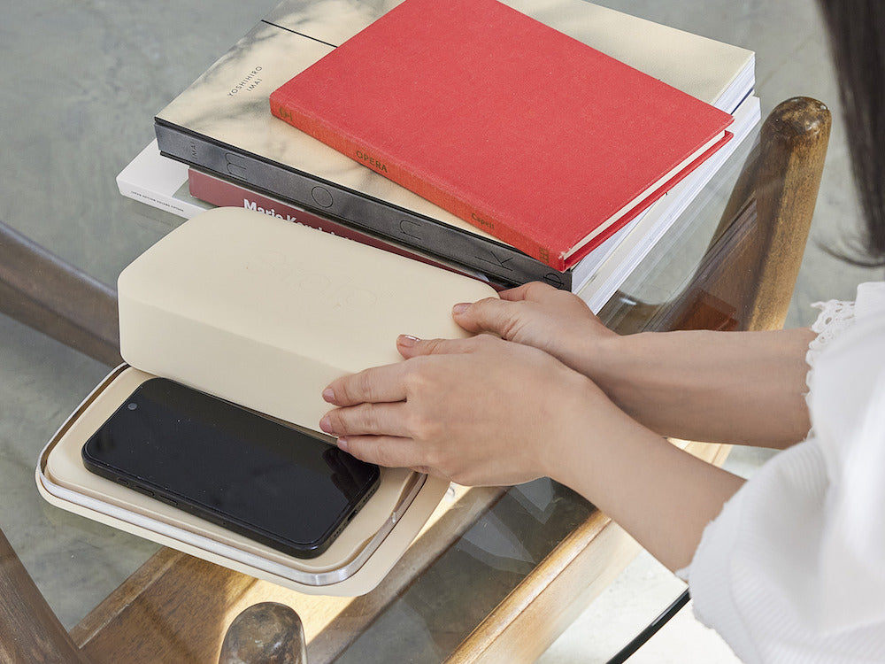 Two hands lowering the Stolp box lid over a phone resting in its tray, on a glass table beside a stack of books